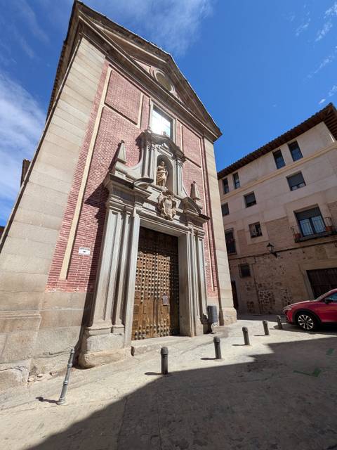 A brick-and-stone church façade with ornate wooden doors and small statue niches under a bright blue sky in Toledo.
