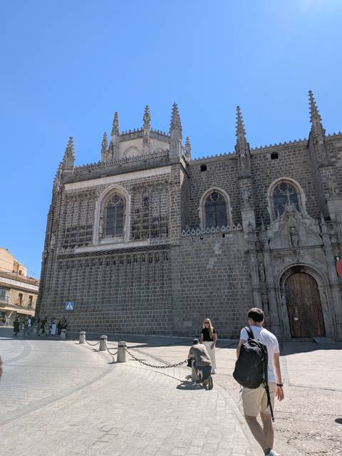 Gothic stone façade of the Monastery of San Juan de los Reyes with tall spires and tourists admiring it below.