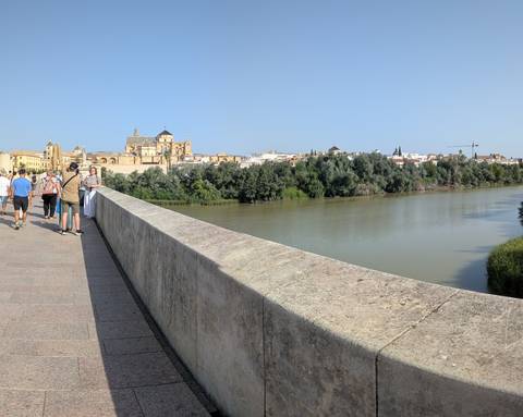 Pedestrians cross Córdoba’s Roman bridge with the Mosque–Cathedral towering over the opposite bank beside the river.