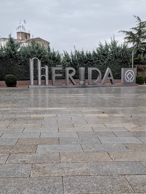 Large metal letters spelling MERIDA on a wet paved plaza with trimmed hedges behind.