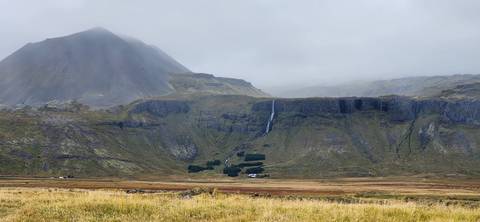 A misty mountainside with a thin waterfall dropping down steep green cliffs under an overcast sky.