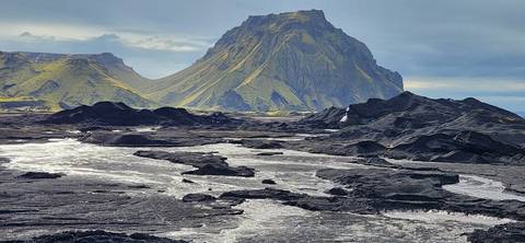 A stark black lava plain with bright streams lies before a rugged green-streaked mountain under a brooding sky.