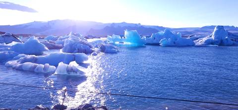 Sparkling sunlight reflects off a lagoon filled with blue icebergs backed by snowy mountains.