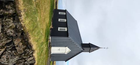 A simple black wooden church stands on a grassy lava field beneath a grey Icelandic sky.