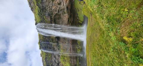 The tall veil of Seljalandsfoss plunges over a mossy cliff into a green meadow under broken clouds.