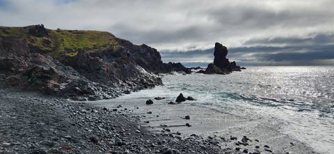 Waves crash against a rugged volcanic shoreline and a lone sea stack under dramatic clouds.