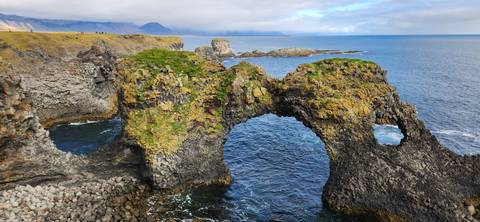 A dramatic natural stone arch covered in moss rises above deep blue Atlantic water along a rugged coast.