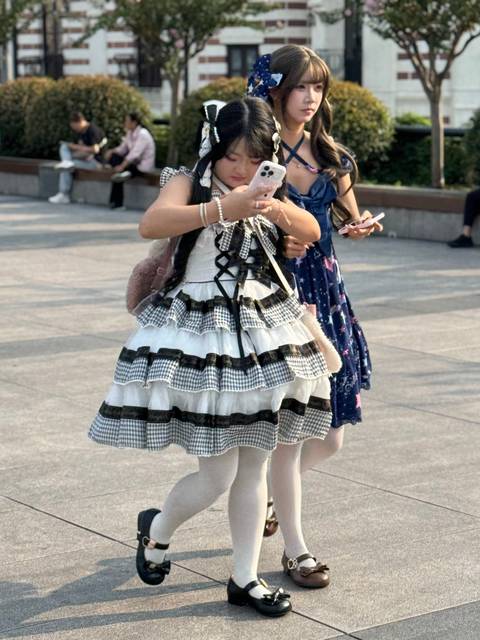 Two young women in frilly, layered dresses check their phones while standing in a sunny public square.