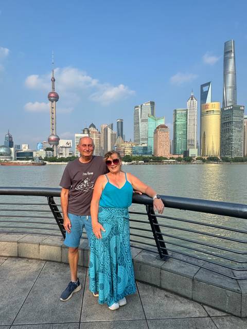 Smiling couple posing on a riverside promenade with Shanghai’s modern skyscrapers across the water under blue skies.