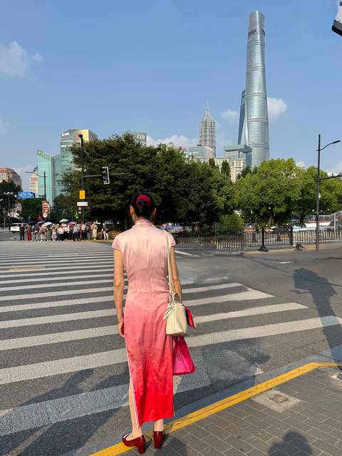 Woman in a pink traditional dress waiting to cross a striped crosswalk with city trees and skyscrapers behind her.