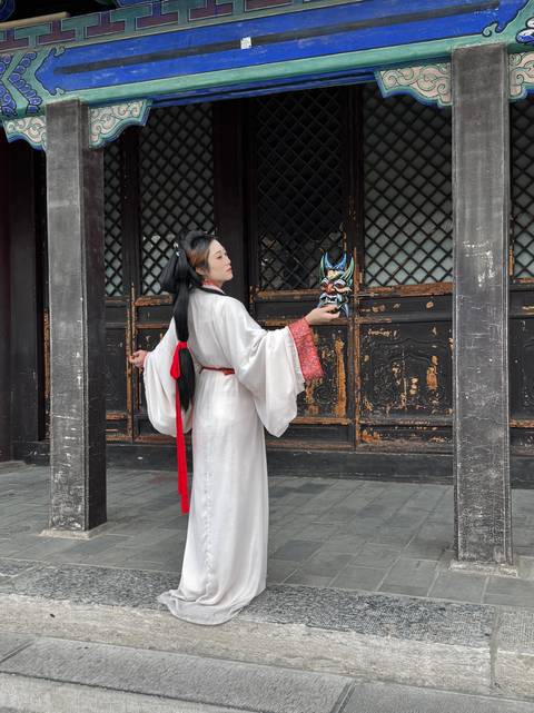 Woman in flowing white traditional costume holding a dramatic opera mask in a historic wooden courtyard.