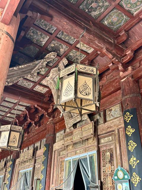 Ornate wooden lantern with intricate Arabic calligraphy hanging beneath richly carved temple eaves.