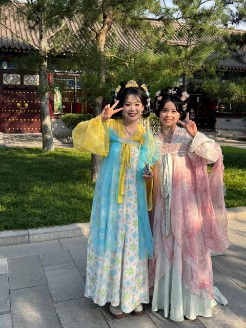 Two young women in flowing traditional dresses flashing peace signs in a sunlit park.