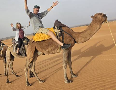 Travelers riding camels across rolling orange dunes of the desert, arms raised in excitement