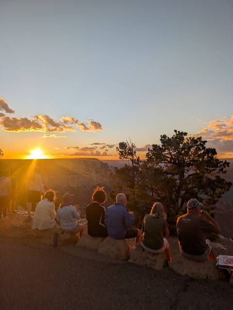 Travellers sit on a canyon rim watching the sun set over rugged cliffs and pinyon pines.