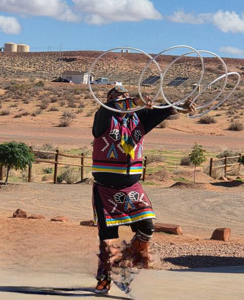 A Navajo hoop dancer performs outdoors in colorful regalia against a desert backdrop.