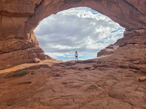 A lone hiker stands beneath a vast natural sandstone arch with dramatic clouds overhead.