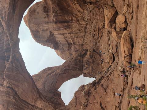Visitors scramble over boulders beneath a pair of intersecting sandstone arches in a rugged canyon.