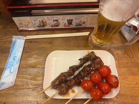 Plate of grilled meat skewers and roasted tomatoes accompanied by a cold beer on a wooden izakaya table.