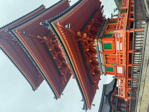 Vivid orange three-tiered pagoda with intricate wooden joinery and green panels against an overcast sky in Kyoto.