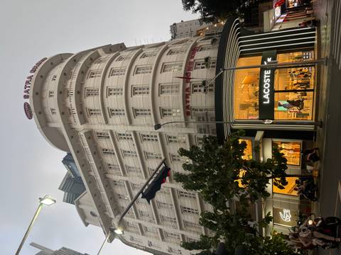 Tall white SATRA Group building with a glowing Lacoste store at street level during dusk in a busy city.