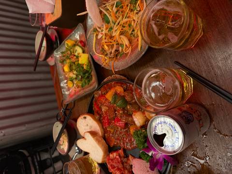 Close-up of Vietnamese dishes: sizzling stew, fresh salads, bread, beer and iced drinks on a rustic table.