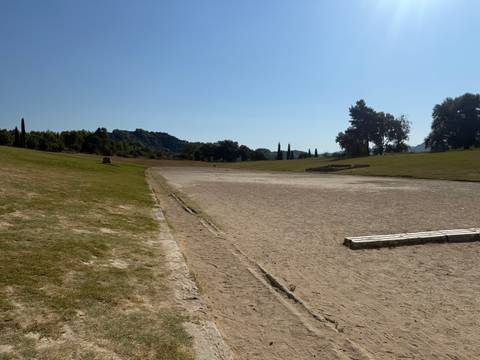 Wide sandy ancient stadium at Olympia surrounded by grassy banks under bright sun.
