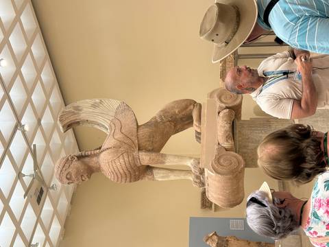 Museum guide speaking to visitors beside a large ancient sphinx statue on a pedestal.