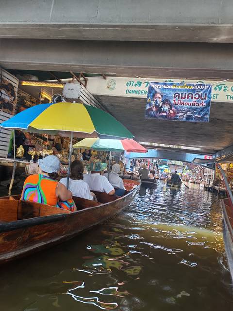 Visitors seated in wooden boats under a low bridge at a Thai floating market surrounded by souvenir stalls