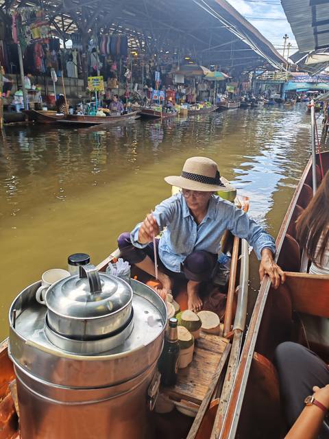 Female vendor preparing food on a small wooden boat at a floating market, stirring ingredients beside another passenger boat