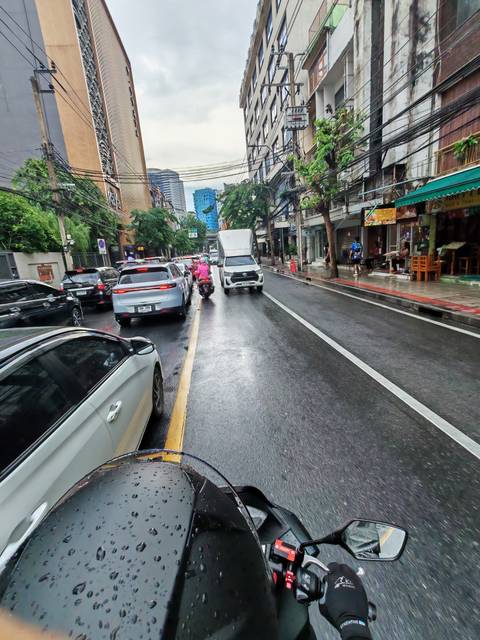 Rain-wet Bangkok city street with cars, motorbikes and pedestrians under gray skies