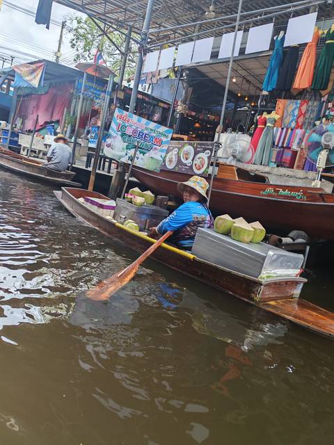 Vendor in blue shirt on small boat selling coconuts at floating market, paddling with wooden oar
