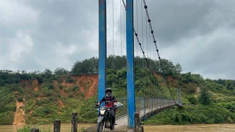 Motorbike rider on dirt bike crossing narrow blue suspension bridge over muddy river