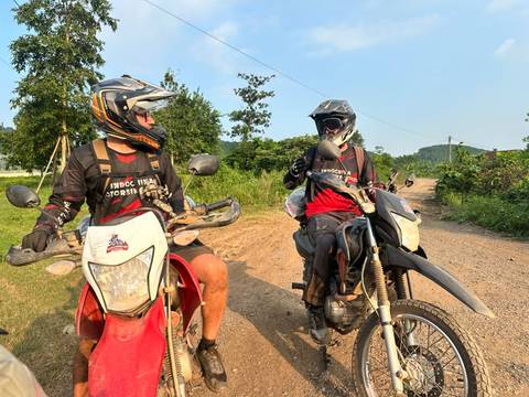 Two dirt bike riders wearing helmets talking on rural track with greenery and blue sky