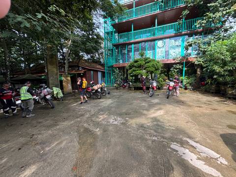 Motorbikes and riders gathered in courtyard of green-glass guesthouse surrounded by trees