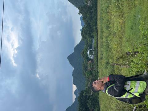 Motorbiker standing in a field smoking, with green peaks and cloudy sky in the background