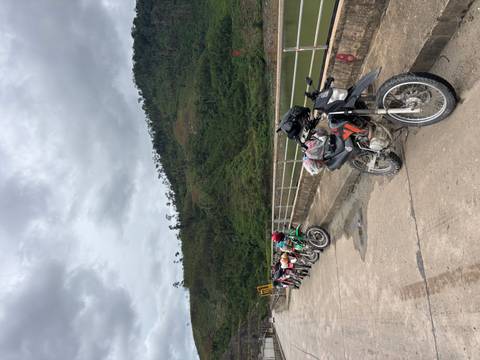 Row of dirt bikes parked on a concrete bridge beside a forested reservoir bank under overcast skies