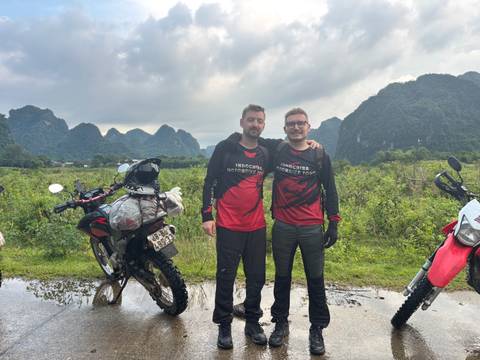 Two motorcyclists in matching jerseys pose with their bikes against limestone karst mountains after rain