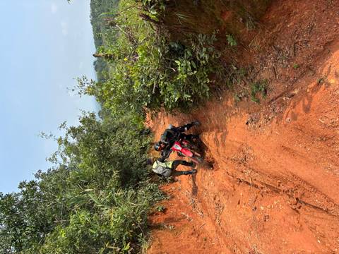 Two riders struggle to push a dirt bike through a narrow red-clay jungle trail
