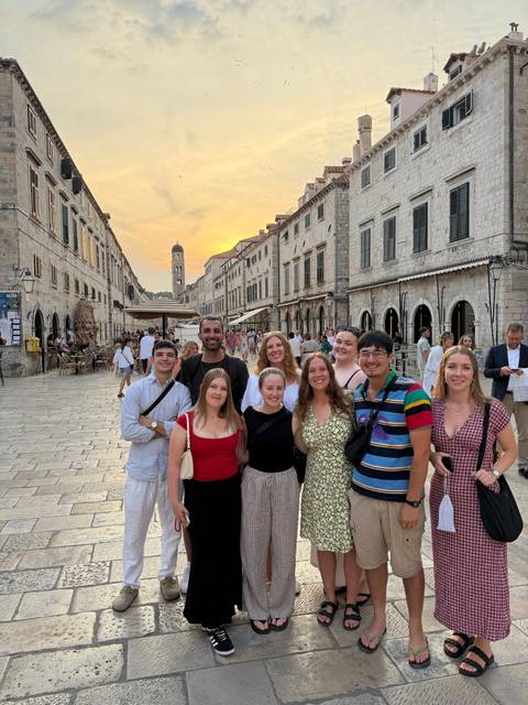Happy tour group poses on Dubrovnik’s historic Stradun at sunset with bell tower and limestone buildings framing the scene.