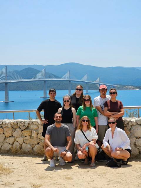 Group poses with Croatia’s Pelješac Bridge and turquoise Adriatic waters behind them on a sunny day.