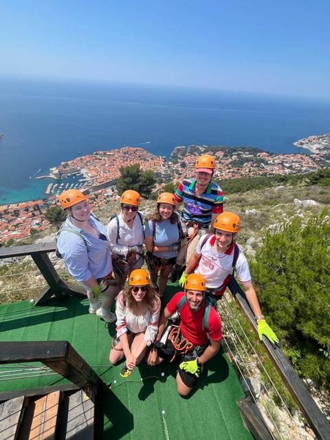 Thrilled travellers in helmets stand on a mountaintop zipline platform with aerial view of Dubrovnik’s old town and sea below.