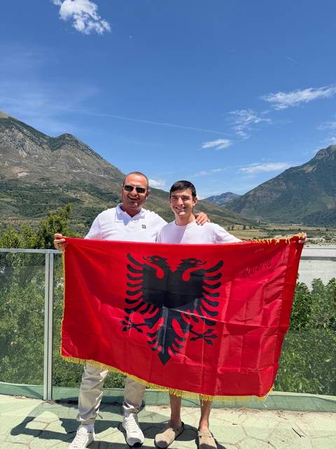 Two travellers proudly display the Albanian flag with rolling green valleys and peaks behind them.