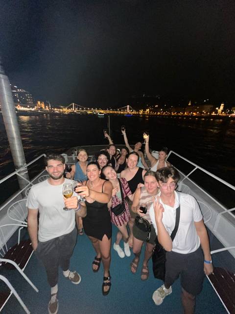 Nighttime selfie of smiling travelers raising drinks on the deck of a river boat with city lights behind.