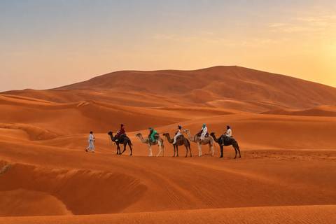 Line of camel riders led by a guide trek across sweeping orange Sahara dunes at sunset.