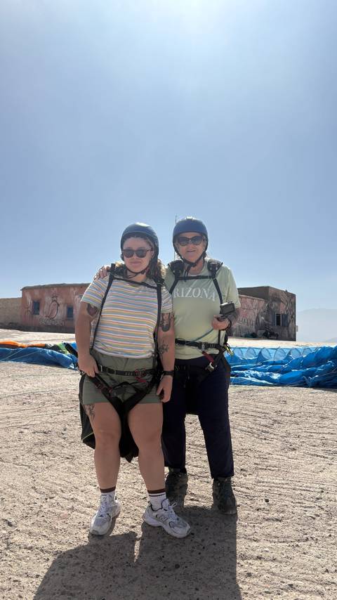 Two women wearing helmets and harnesses prepare for paragliding beside blue canopies on a plateau.