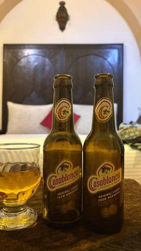 Two Moroccan beer bottles and a glass placed on a hotel bed with cushions in background.