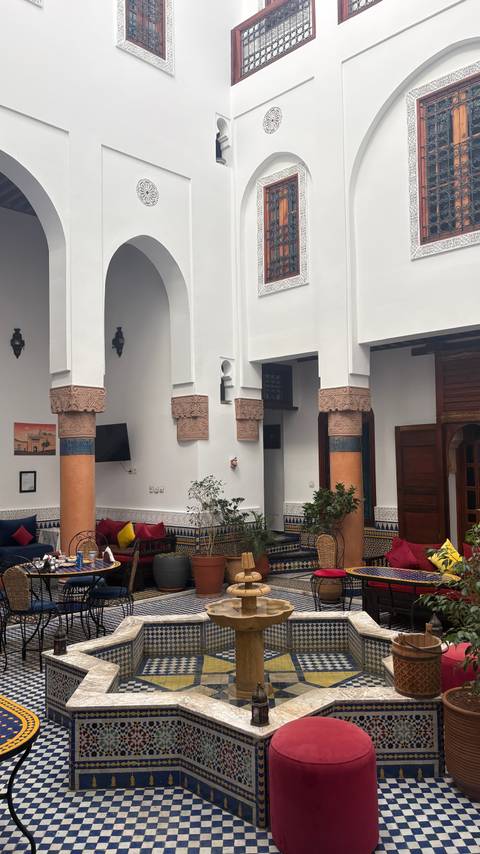Traditional riad courtyard with carved columns, tiled walls and colourful cushions around a fountain.