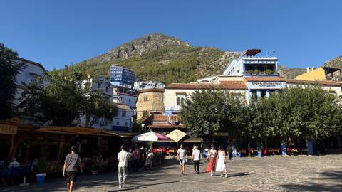Sunny plaza framed by white-and-blue buildings with a mountain backdrop where locals and tourists stroll.