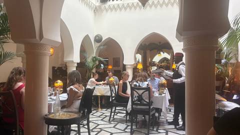 Elegant restaurant atrium with white arches where diners enjoy meals served by a waiter in traditional vest and fez.